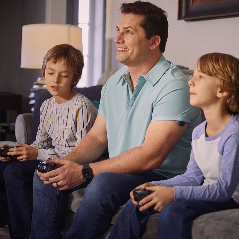 Uomo che gioca ai videogiochi con i figli mentre indossa un CGM impiantabile sul braccio.