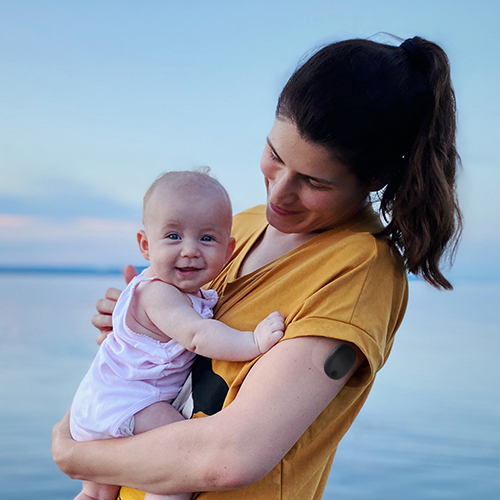 Une femme à la plage avec un CGM de longue durée Eversense et portant son bébé.